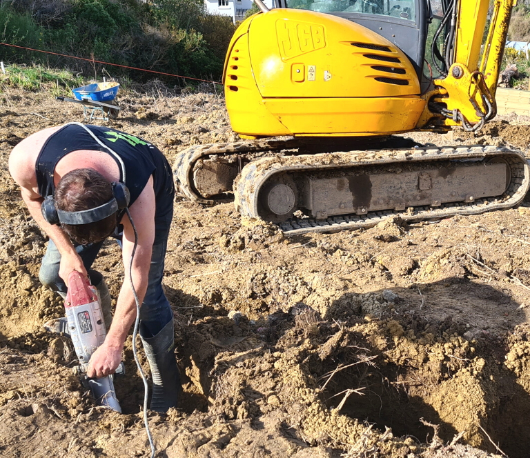 Builder digging hole for foundation supports, with yellow digger in the background and muddy field around him